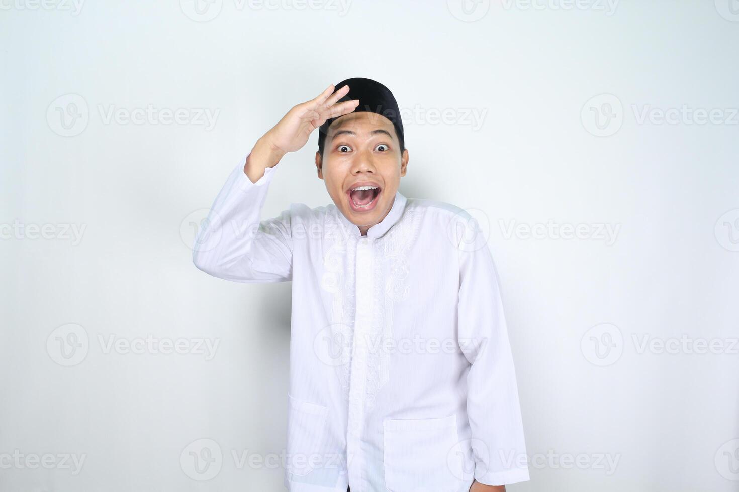 funny muslim man asian show surprised expression while looking far away at camera with hand raised over his head isolated on white background photo