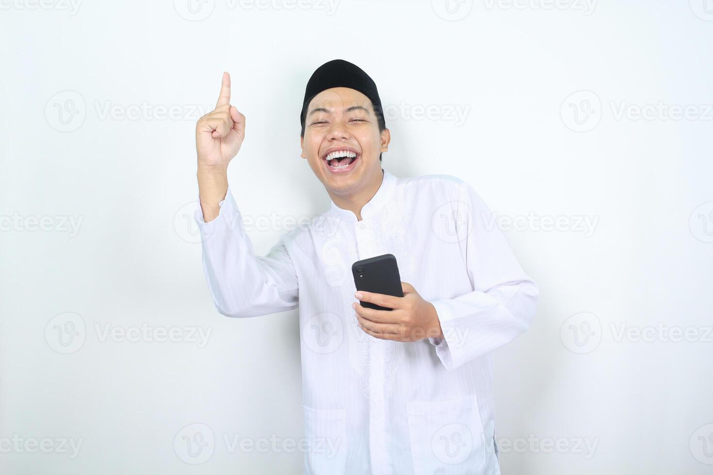 ecstatic asian muslim man laughing while holding phone with pointing finger upward isolated photo