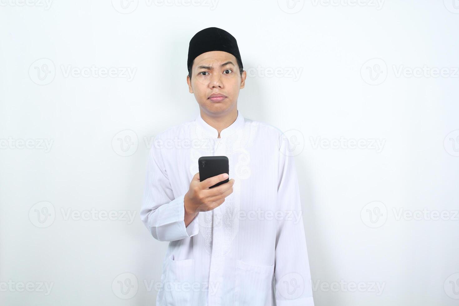 confused muslim asian man holding mobile phone looking at camera isolated on white background photo