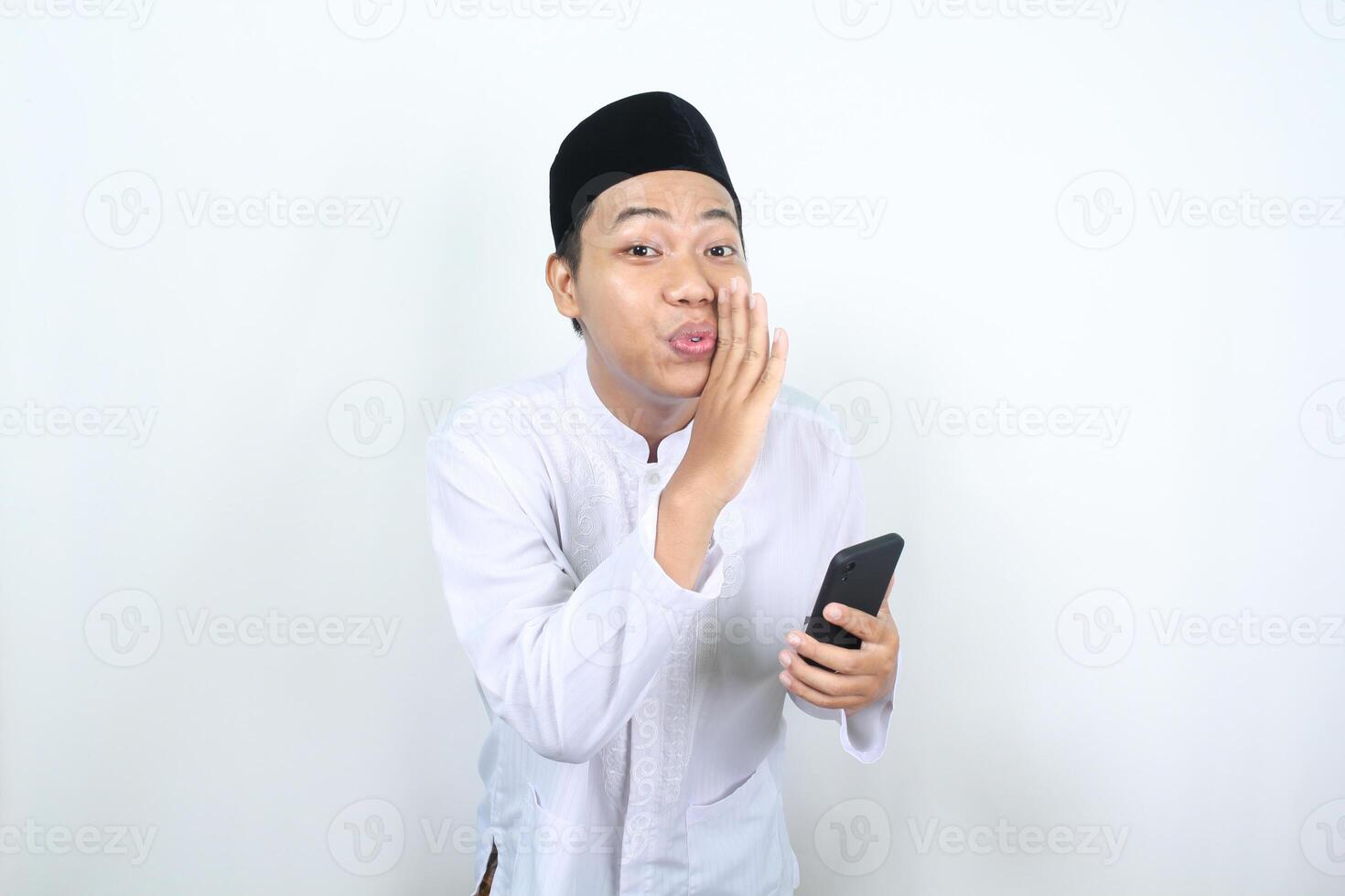 delighted asian muslim man do whispering gesture with holding phone isolated photo