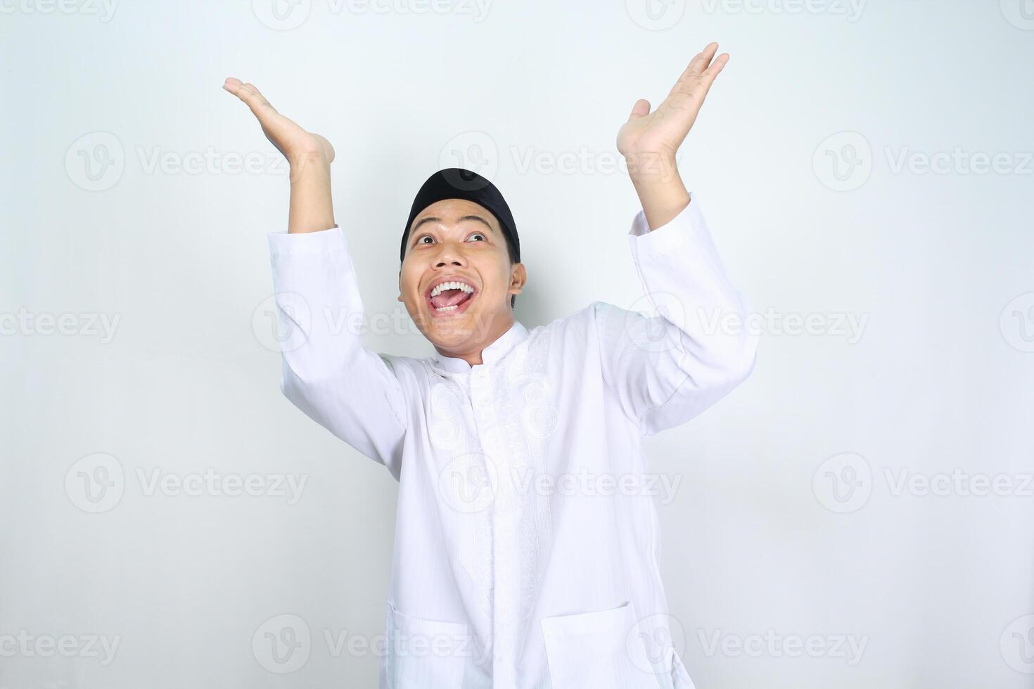 funny asian muslim man looking up with surprised expression and raising hand to presenting isolated on white background photo