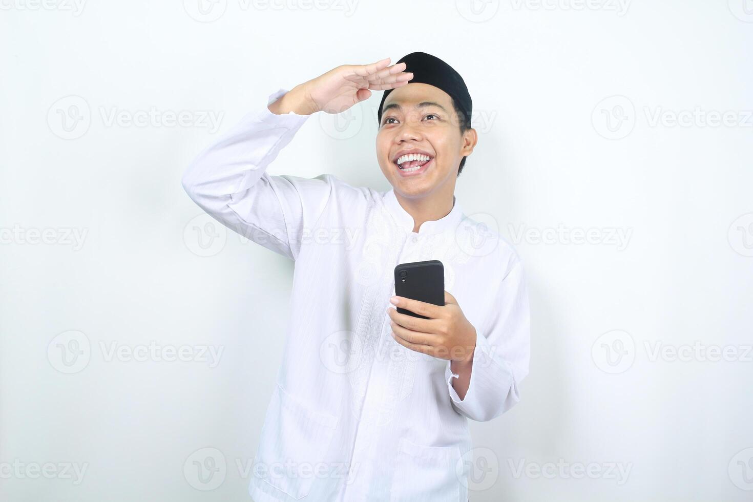 smiling asian muslim man raising hand over head show looking far away gesture with holding mobile phone isolated photo