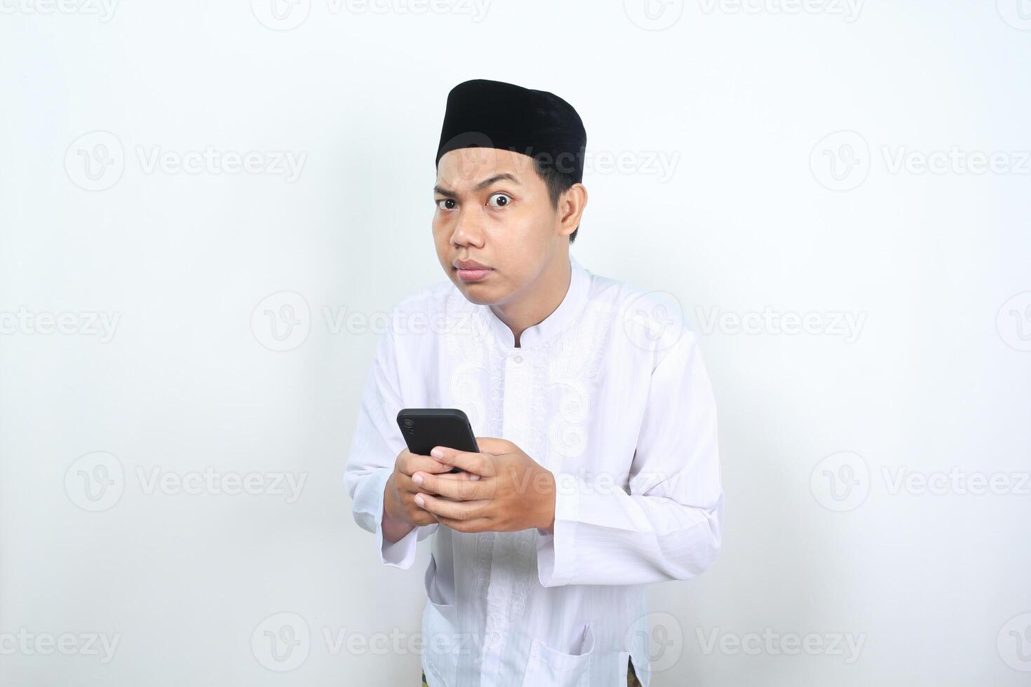 closeup of muslim asian man holding mobile phone looking at camera with confused expression isolated on white background photo