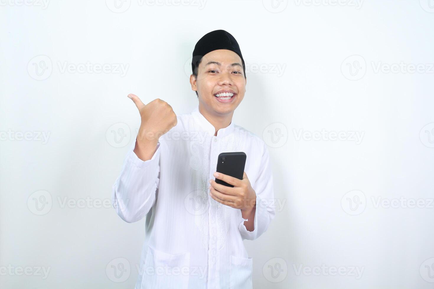 smiling asian muslim man holding phone with pointing to beside isolated photo