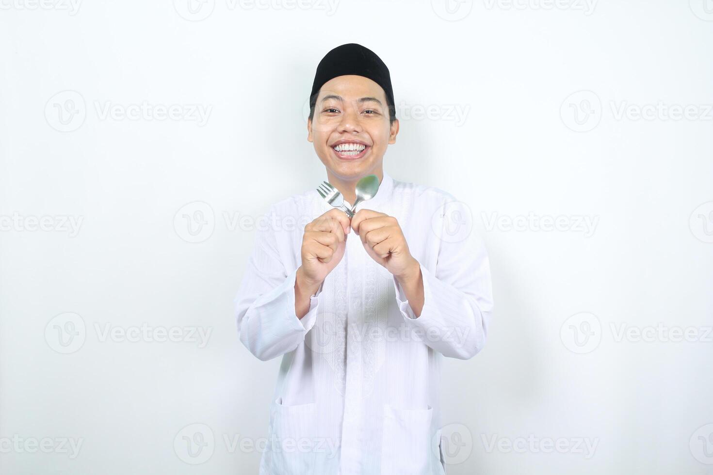 smiling muslim asian man holding crossed spoon and fork waiting for break fasting in ramadhan isolated photo