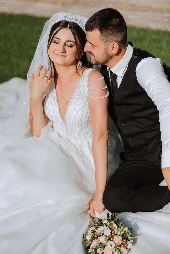 Beautiful couple sitting and hugging outdoors on their wedding day, relaxing on the best summer day. A handsome groom and an elegant bride in a magnificent wedding dress. photo