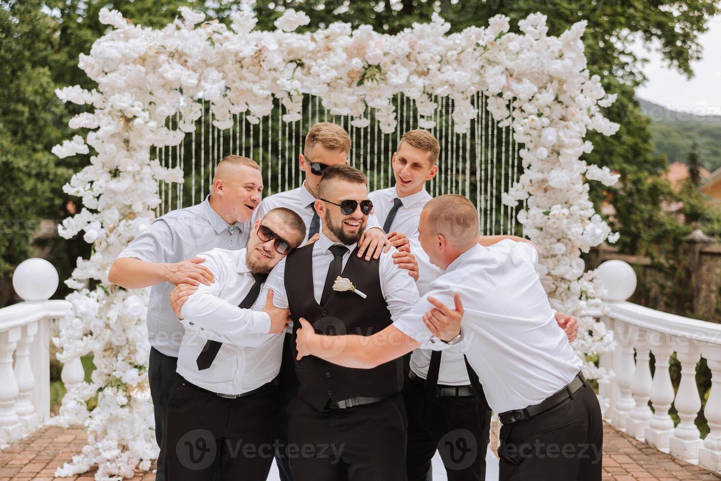 Cheerful, young, energetic witnesses of the groom next to the groom. Friends congratulate the groom. The groom in a vest and his friends in white shirts pose near the wedding arch. Wedding in nature. photo