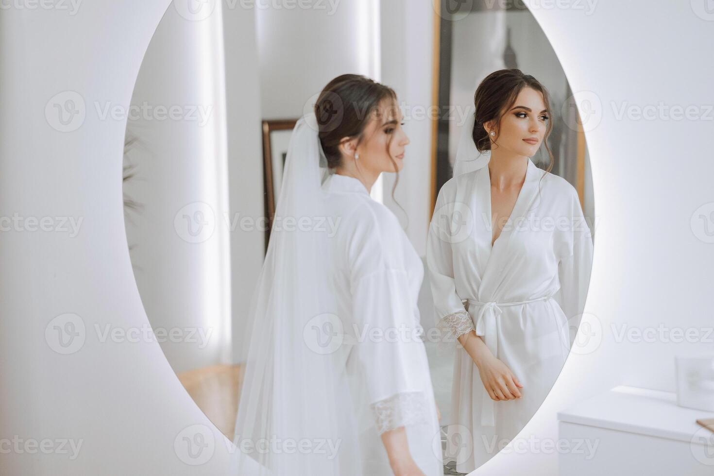Bride in a wedding dress looking in the mirror. Beautiful young girl bride in the studio near the mirror in a white robe. photo