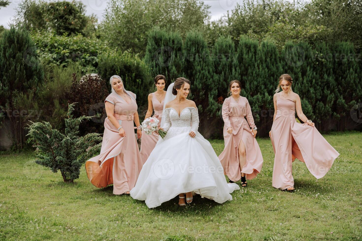 A brunette bride and her bridesmaids in identical pink dresses are walking against the background of nature. Girls in identical dresses are making out at a wedding. Wedding in nature. photo