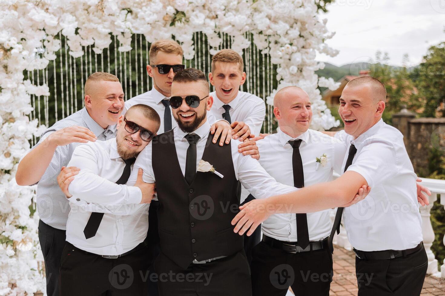 Cheerful, young, energetic witnesses of the groom next to the groom. Friends congratulate the groom. The groom in a vest and his friends in white shirts pose near the wedding arch. Wedding in nature. photo
