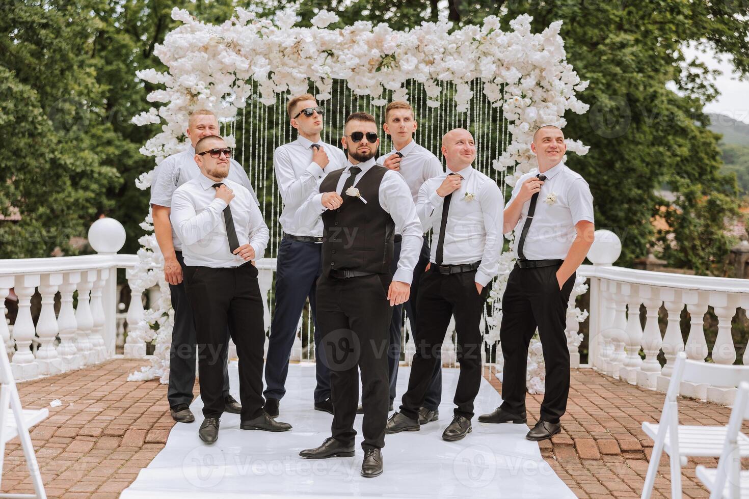 Cheerful, young, energetic witnesses of the groom next to the groom. Friends congratulate the groom. The groom in a vest and his friends in white shirts pose near the wedding arch. Wedding in nature. photo