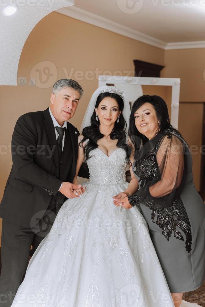 Happy parents hold the hands of the bride who is getting married and support her, wearing a wedding dress and a long veil. photo