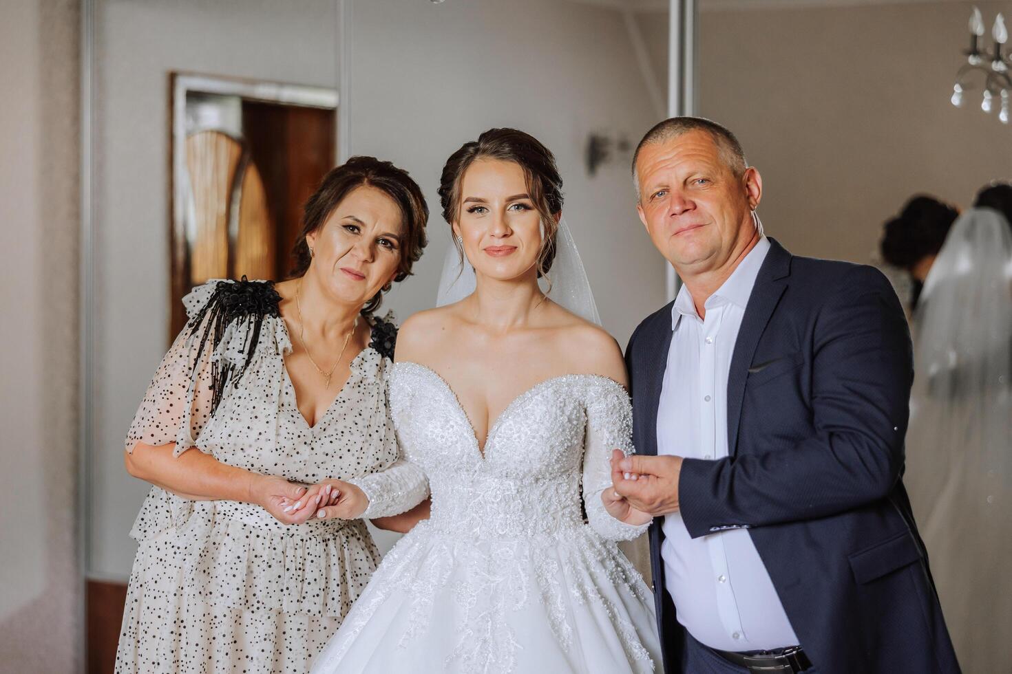 Happy parents hold the hands of the bride who is getting married and support her, wearing a wedding dress and a long veil. photo