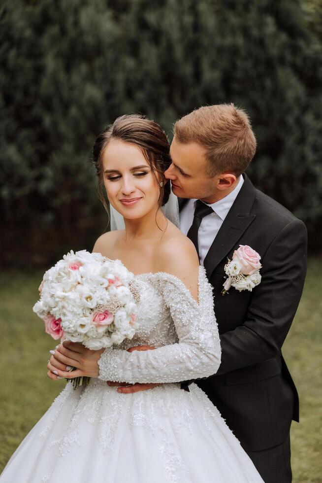 A handsome groom embraces his bride in a lush white dress and smiles in a beautiful outdoor setting. Under the open sky. High quality photo. A newlywed couple poses together on a sunny summer day. photo