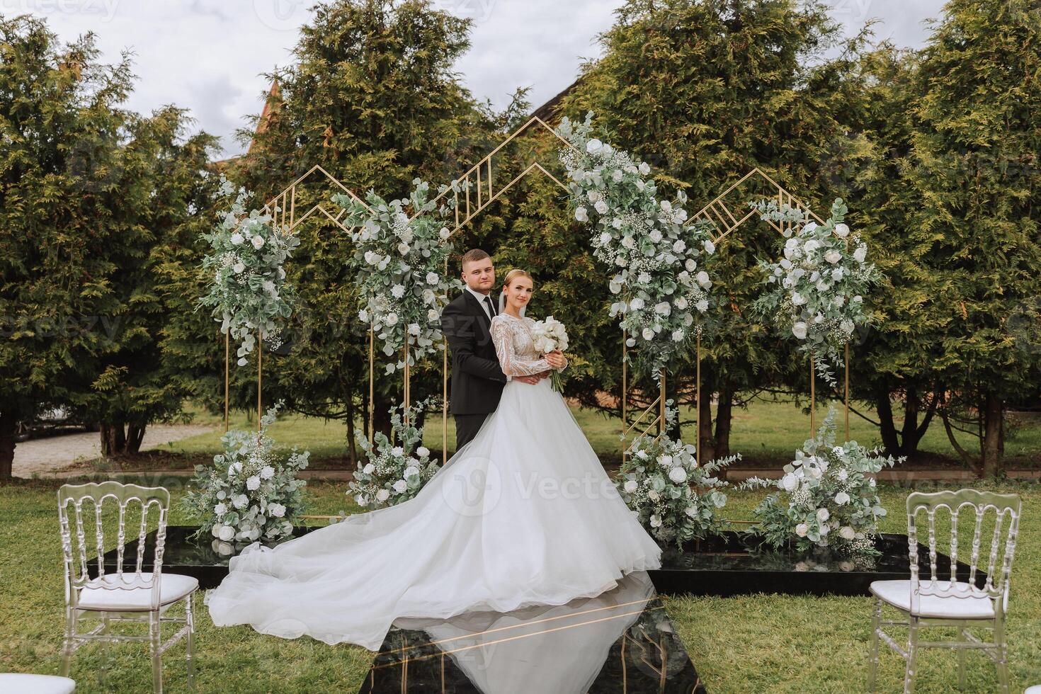 A handsome groom embraces his bride in a lush white dress and smiles in a beautiful outdoor setting. Under the open sky. High quality photo. A newlywed couple poses together on a sunny summer day. photo