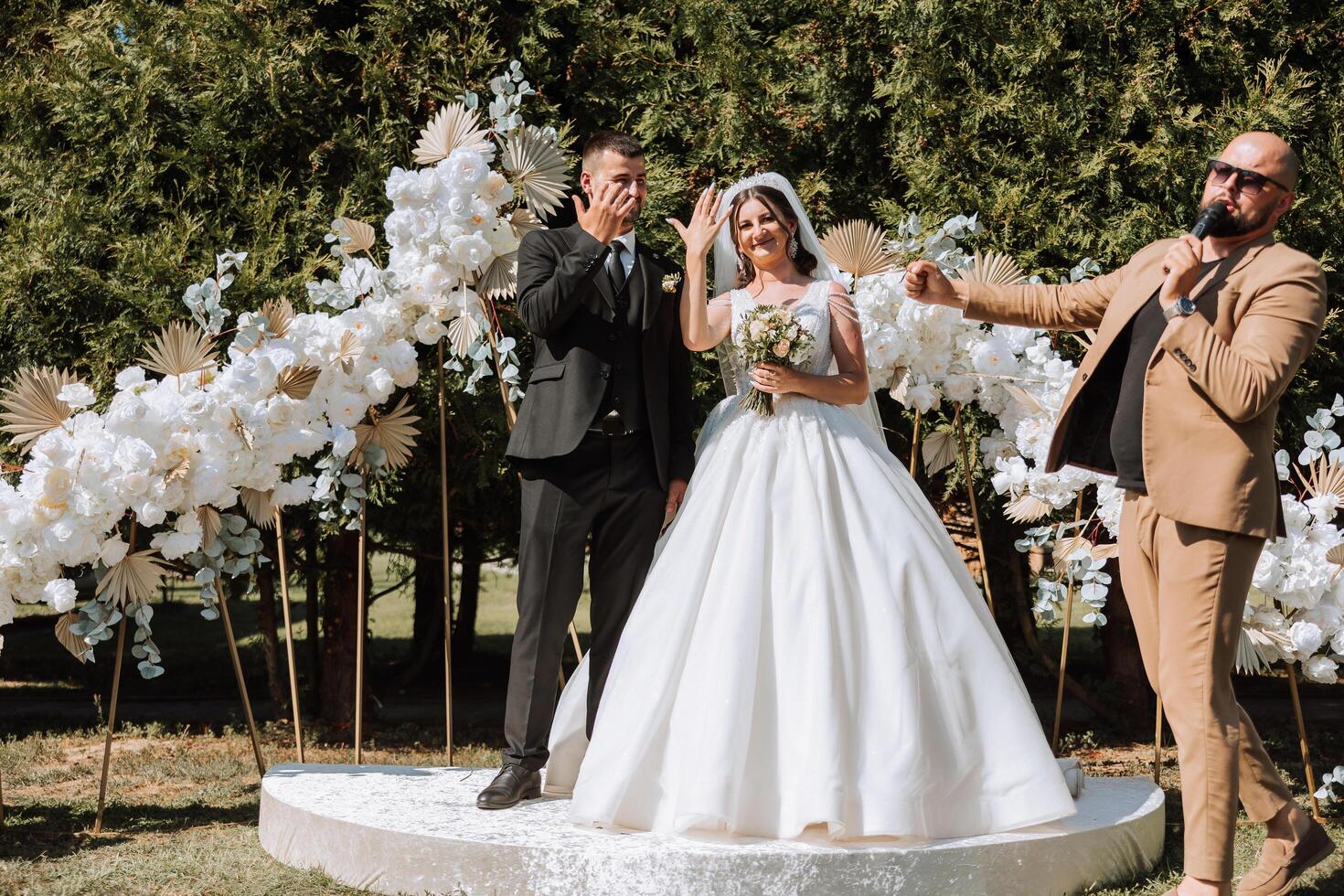 Wedding ceremony in nature. The bride and groom near the flower arch. Master of ceremonies in dark glasses at a wedding during a performance against the background of the bride and groom. photo