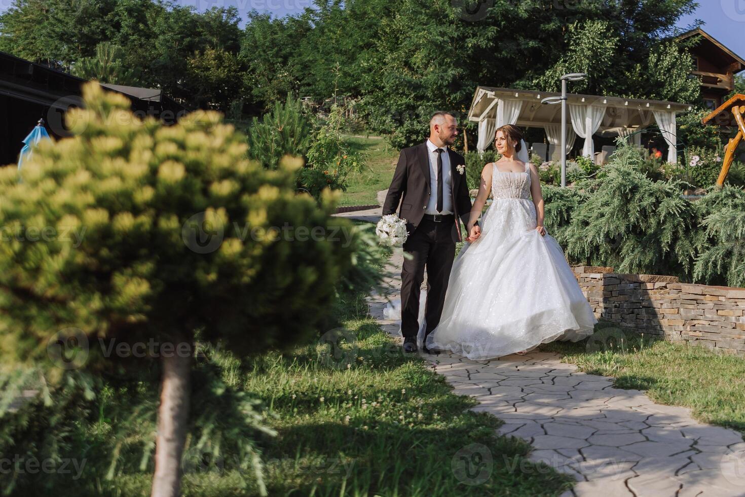 A handsome groom and an elegant bride in a lush white dress are walking in a summer park. Happy bride and groom getting ready for their best day. photo