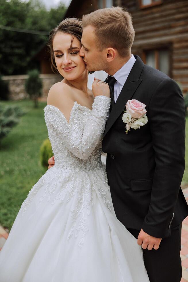 A handsome groom embraces his bride in a lush white dress and smiles in a beautiful outdoor setting. Under the open sky. High quality photo. A newlywed couple poses together on a sunny summer day. photo
