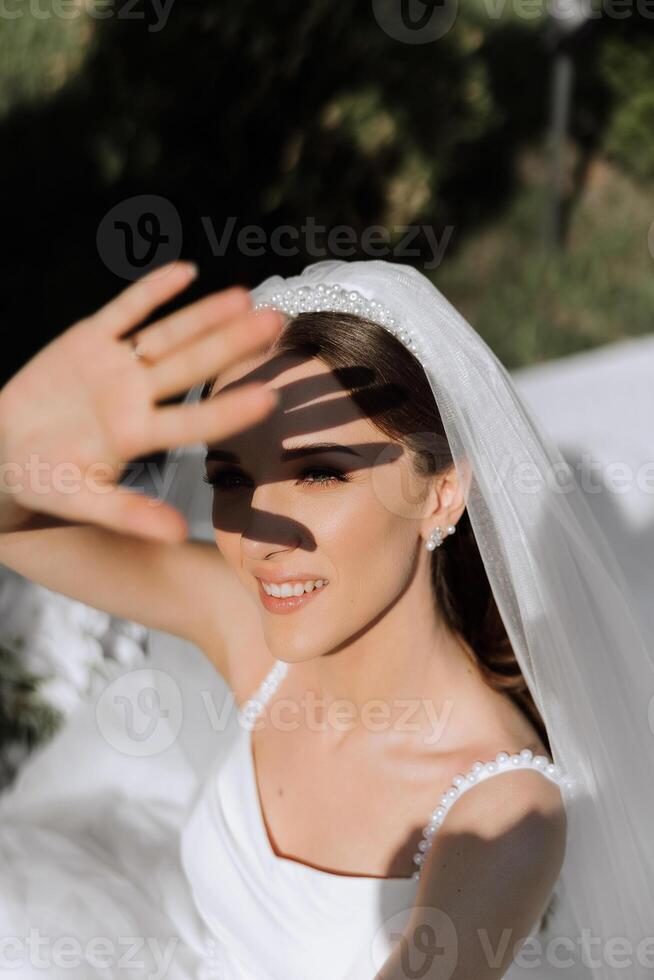 un hermosa novia en un blanco vestir cubre su cara con su mano, proteger su ojos desde el del sol rayos verano boda. foto