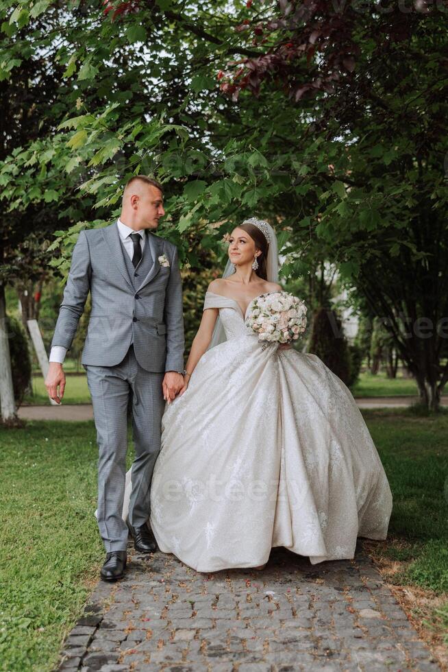 A handsome groom and an elegant bride in a lush white dress are walking in a summer park. Happy bride and groom getting ready for their best day. photo