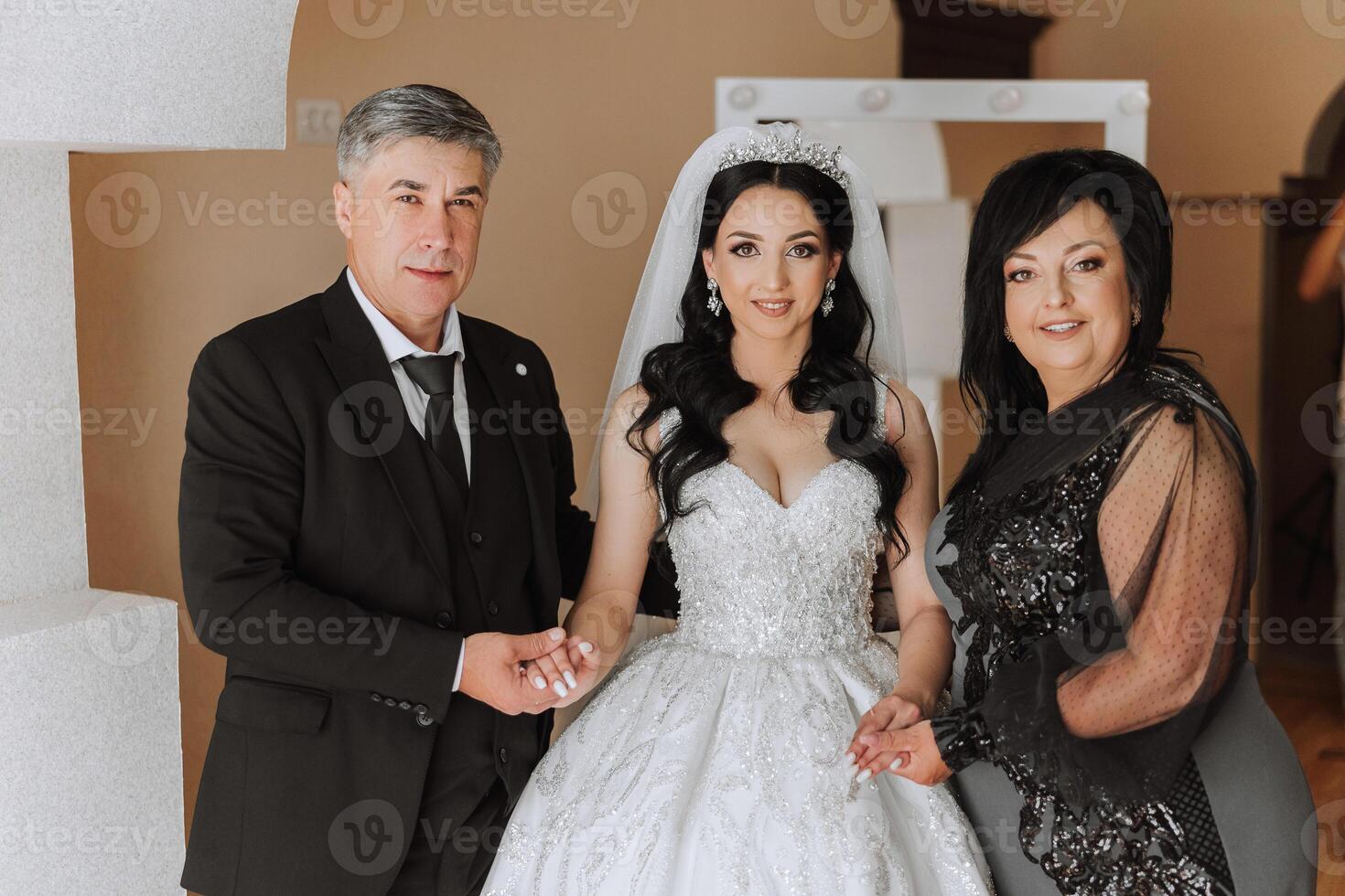 Happy parents hold the hands of the bride who is getting married and support her, wearing a wedding dress and a long veil. photo