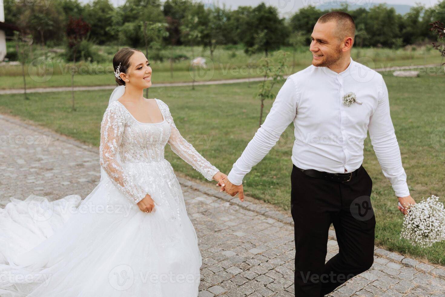 A handsome groom and an elegant bride in a lush white dress are walking in a summer park. Happy bride and groom getting ready for their best day. photo