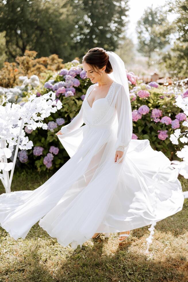 un hermosa morena novia en un elegante túnica es caminando en un parque con verde césped y rosado hortensia flores mañana, reuniones, Boda retrato de un linda muchacha. fotografía y concepto. verano. foto