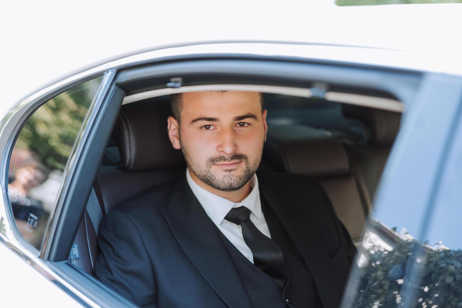 a handsome groom sits with a wedding bouquet inside a white car and looks out the window. A happy moment on the wedding day. photo