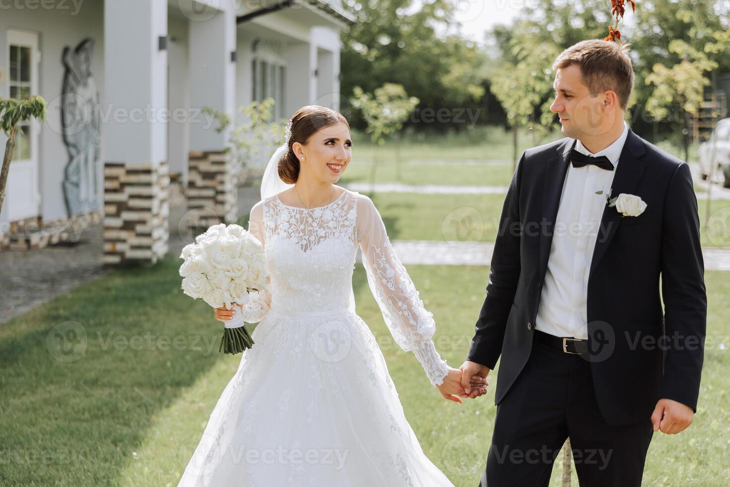 A handsome groom and an elegant bride in a lush white dress are walking in a summer park. Happy bride and groom getting ready for their best day. photo