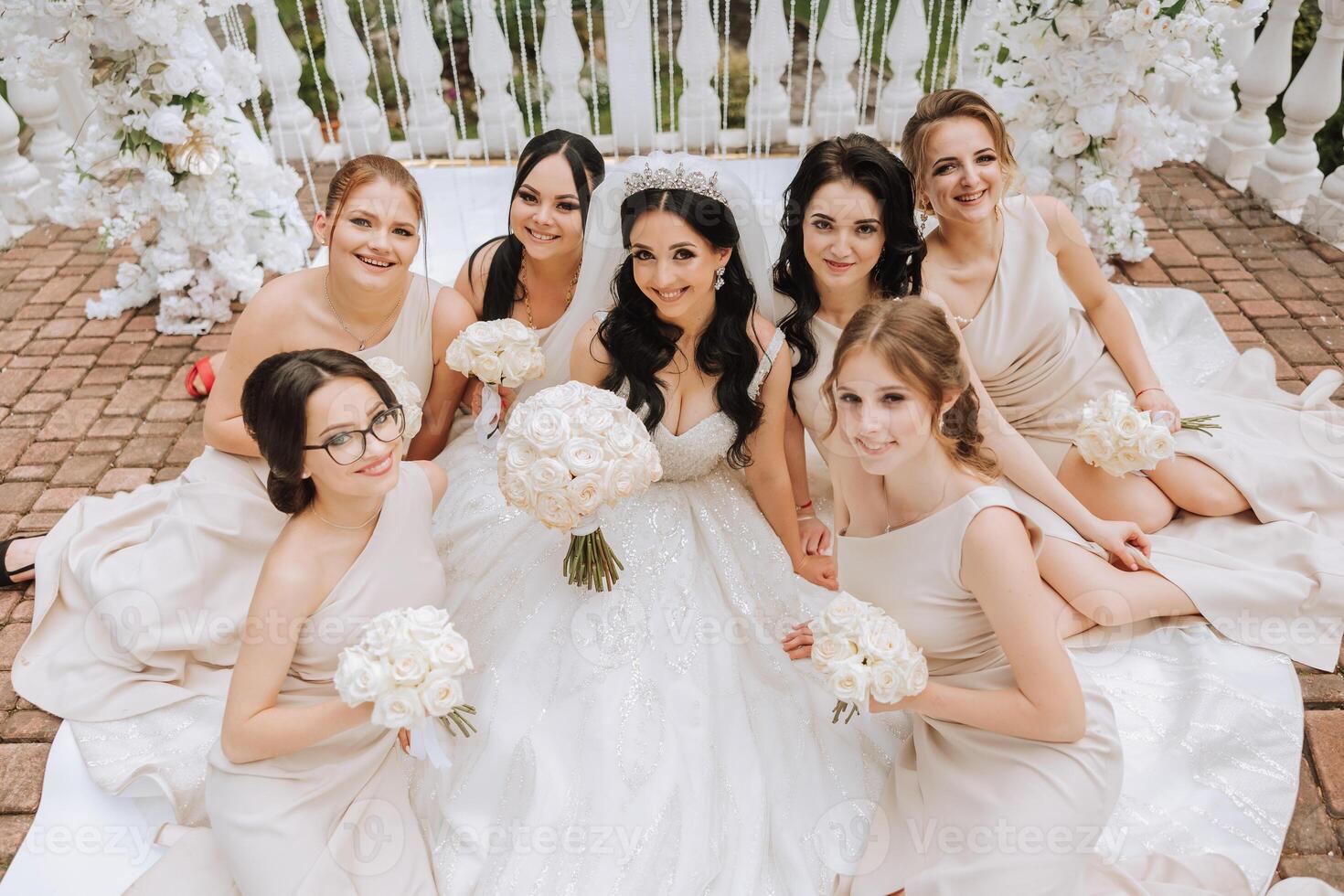 A brunette bride and her bridesmaids in matching cream dresses sit with the bride and rejoice with flowers in their hands near the ceremonial arch. Wedding in nature. photo