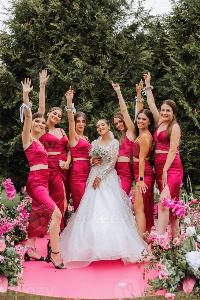 A brunette bride and her bridesmaids in matching pink dresses are standing near the solemn arch. Girls in identical dresses are making out at a wedding. Wedding in nature. photo