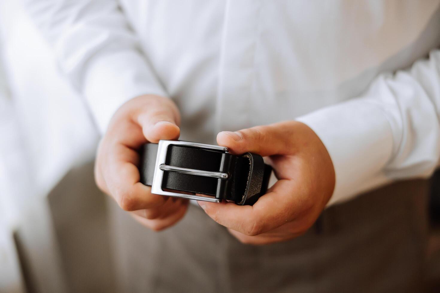 de cerca de un hombre participación un negro cuero cinturón. un hombre en un blanco camisa, sin un rostro, sostiene un negro cuero cinturón en su manos. el Mañana de el novio a el boda. foto