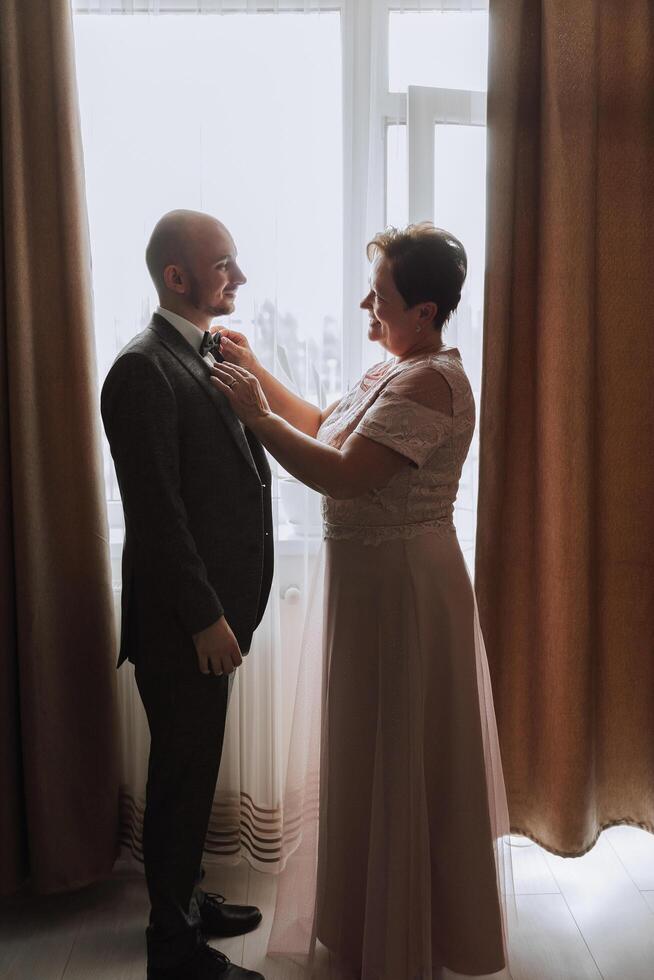 mother helps her adult son prepare for the wedding ceremony. An emotional and touching moment at a wedding. A mother hugs her son photo