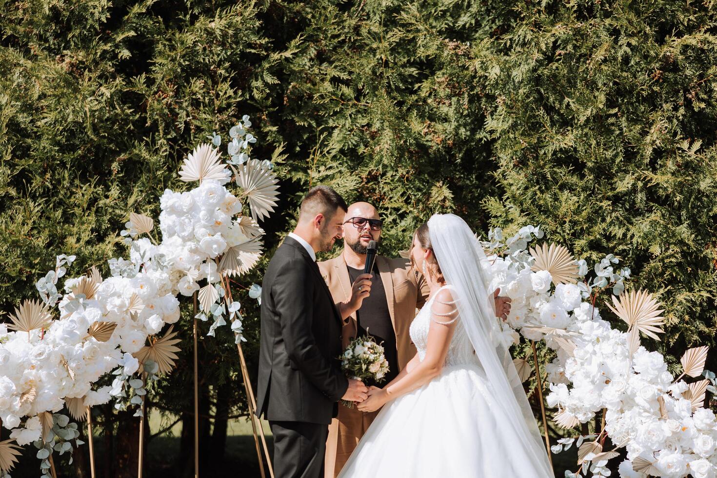 Boda ceremonia en naturaleza. el novia y novio cerca el flor arco. Maestro de ceremonias en oscuro lentes a un Boda durante un actuación en contra el antecedentes de el novia y novio. foto