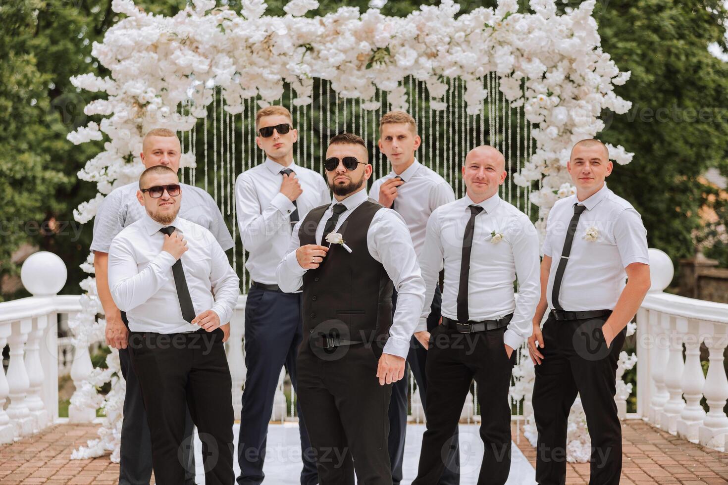 Cheerful, young, energetic witnesses of the groom next to the groom. Friends congratulate the groom. The groom in a vest and his friends in white shirts pose near the wedding arch. Wedding in nature. photo