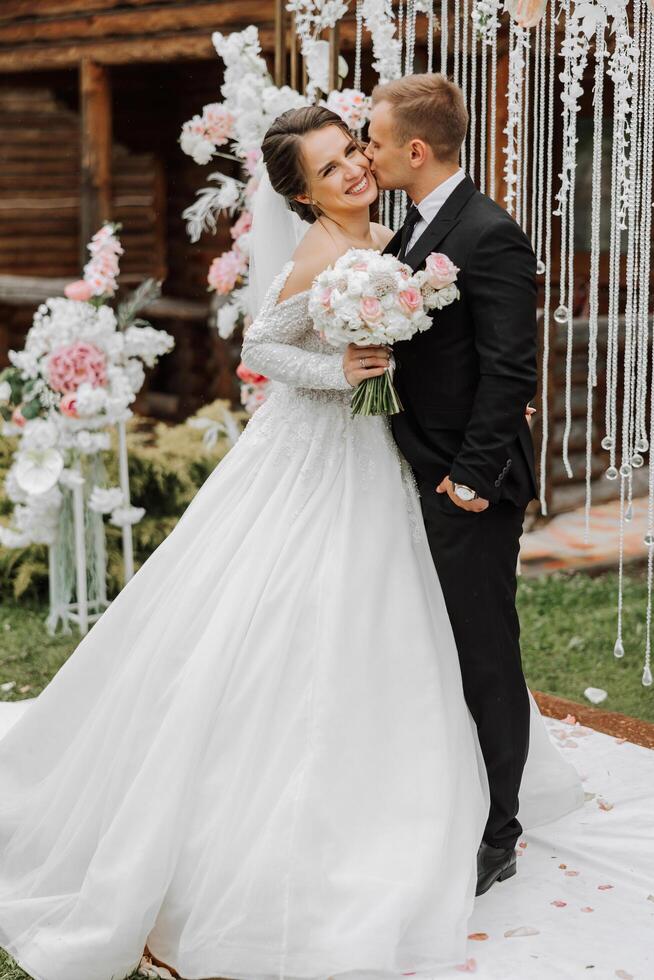 A handsome groom embraces his bride in a lush white dress and smiles in a beautiful outdoor setting. Under the open sky. High quality photo. A newlywed couple poses together on a sunny summer day. photo