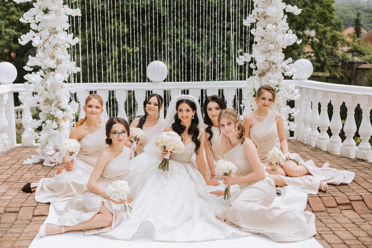 A brunette bride and her bridesmaids in matching cream dresses sit with the bride and rejoice with flowers in their hands near the ceremonial arch. Wedding in nature. photo