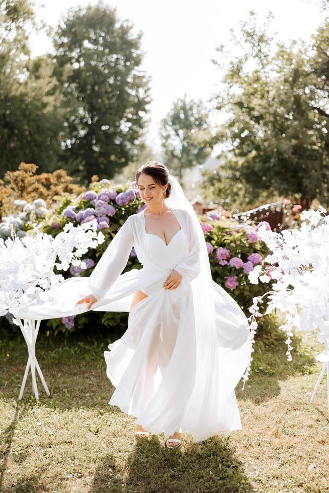 un hermosa morena novia en un elegante túnica es caminando en un parque con verde césped y rosado hortensia flores mañana, reuniones, Boda retrato de un linda muchacha. fotografía y concepto. verano. foto