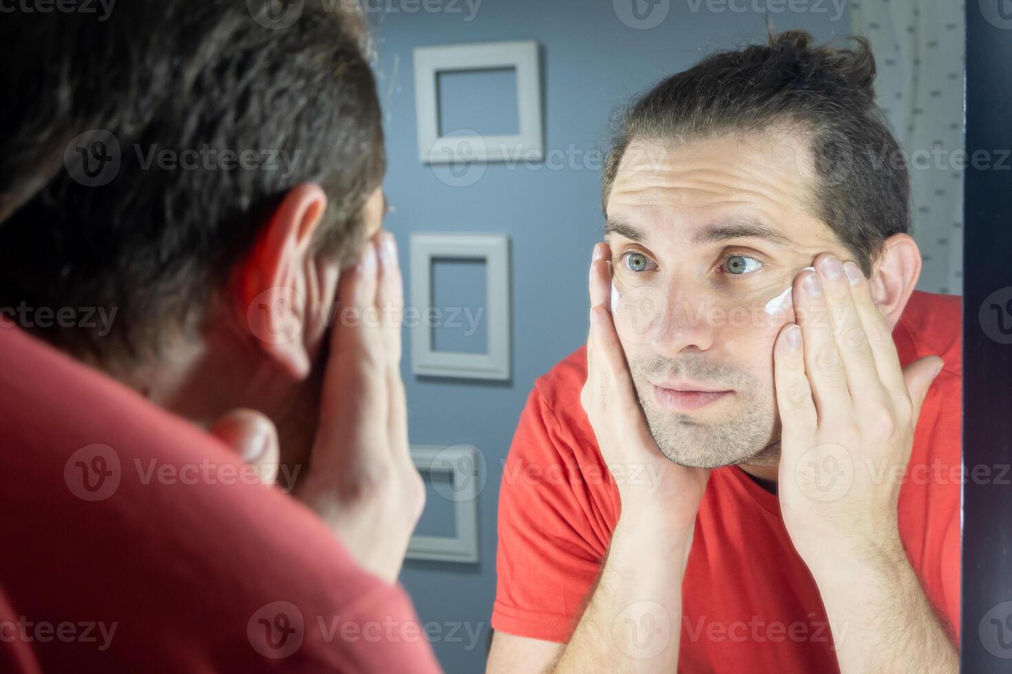 piel cuidado hombre. hombre aplica crema a su cara en frente de el espejo. diario piel cuidado para un hombre. natural foto a hogar.