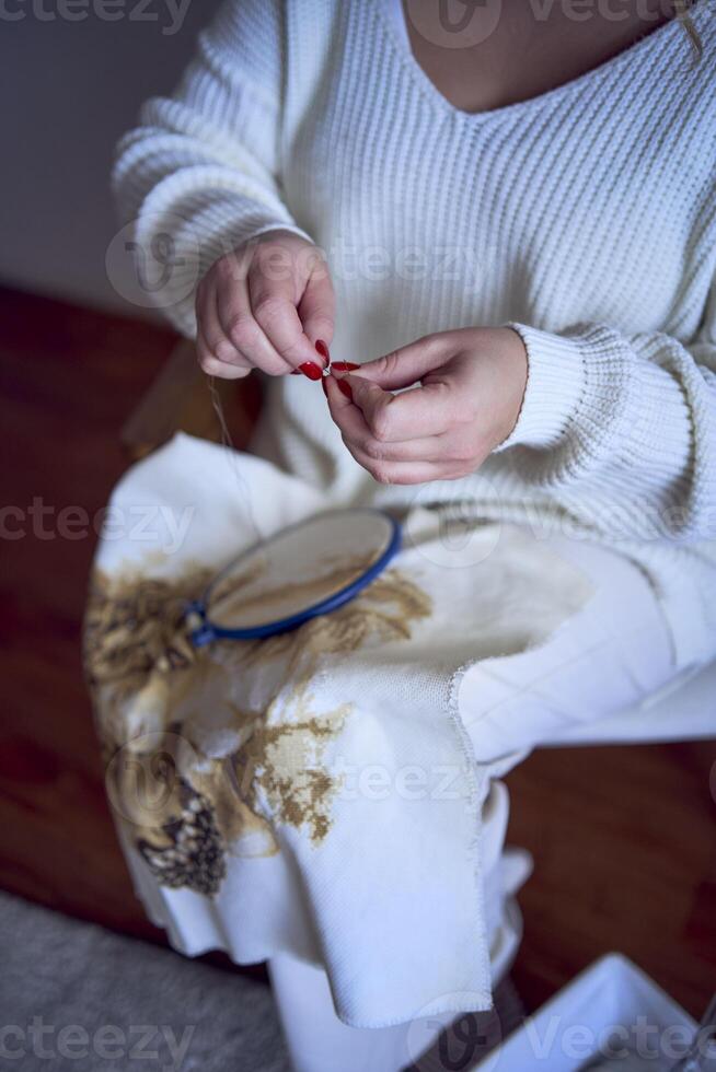 midsize woman in light clothes embroiders a picture in gold and beige tones while sitting on a white chair in a light room photo