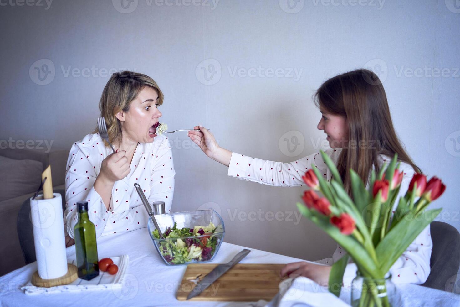 a teenage girl and her mother in pajamas are cooking and eating a fresh green and tomato salad together photo