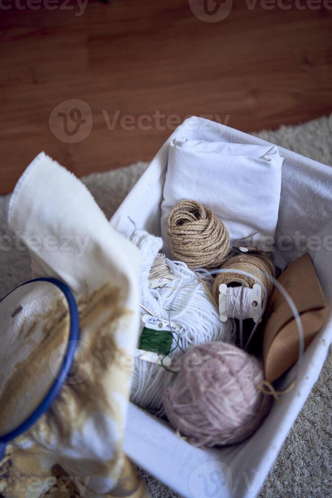 midsize woman in light clothes embroiders a picture in gold and beige tones while sitting on a white chair in a light room photo