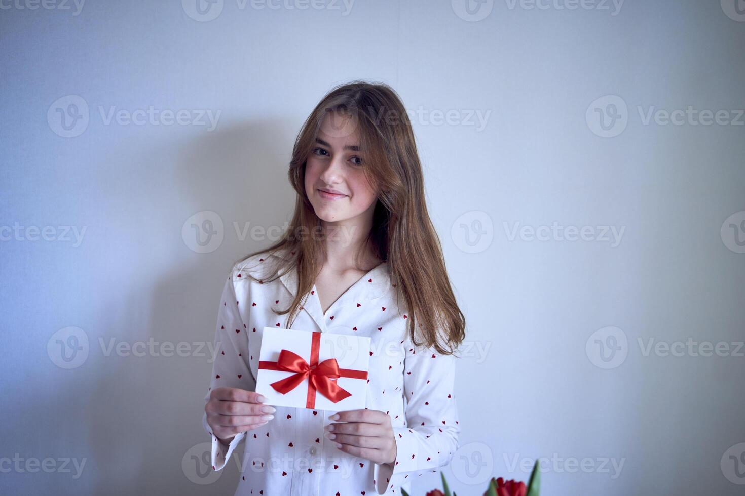 un regalo certificado en el manos de un Adolescente niña vistiendo blanco pijama con rojo corazones foto