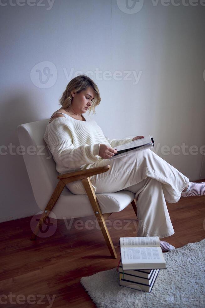 a medium-sized woman in light clothes reads a book while sitting in a white chair in a light room photo