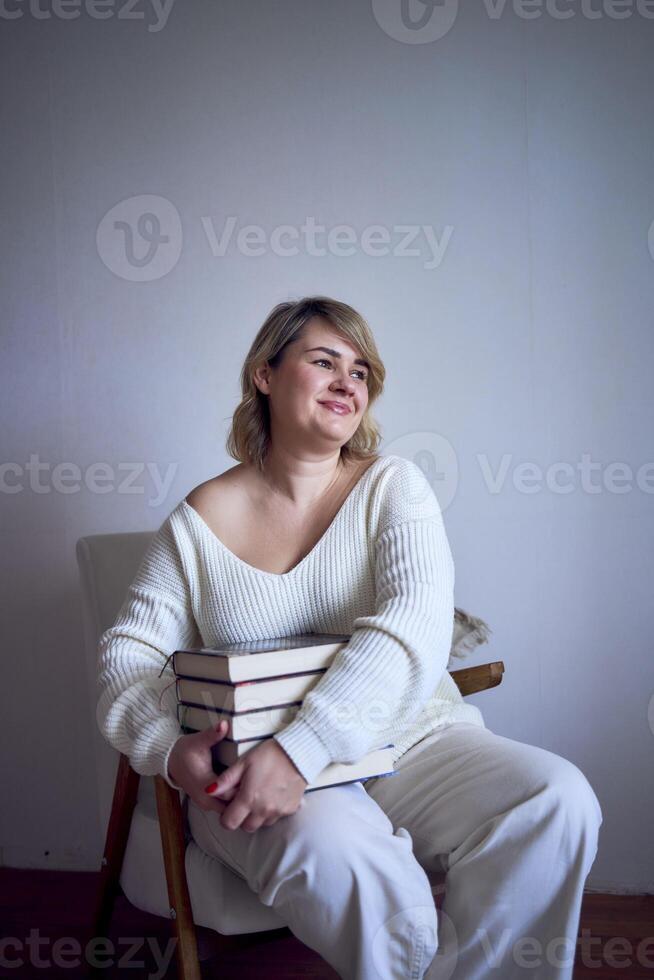 a medium-sized woman in light clothes reads a book while sitting in a white chair in a light room photo