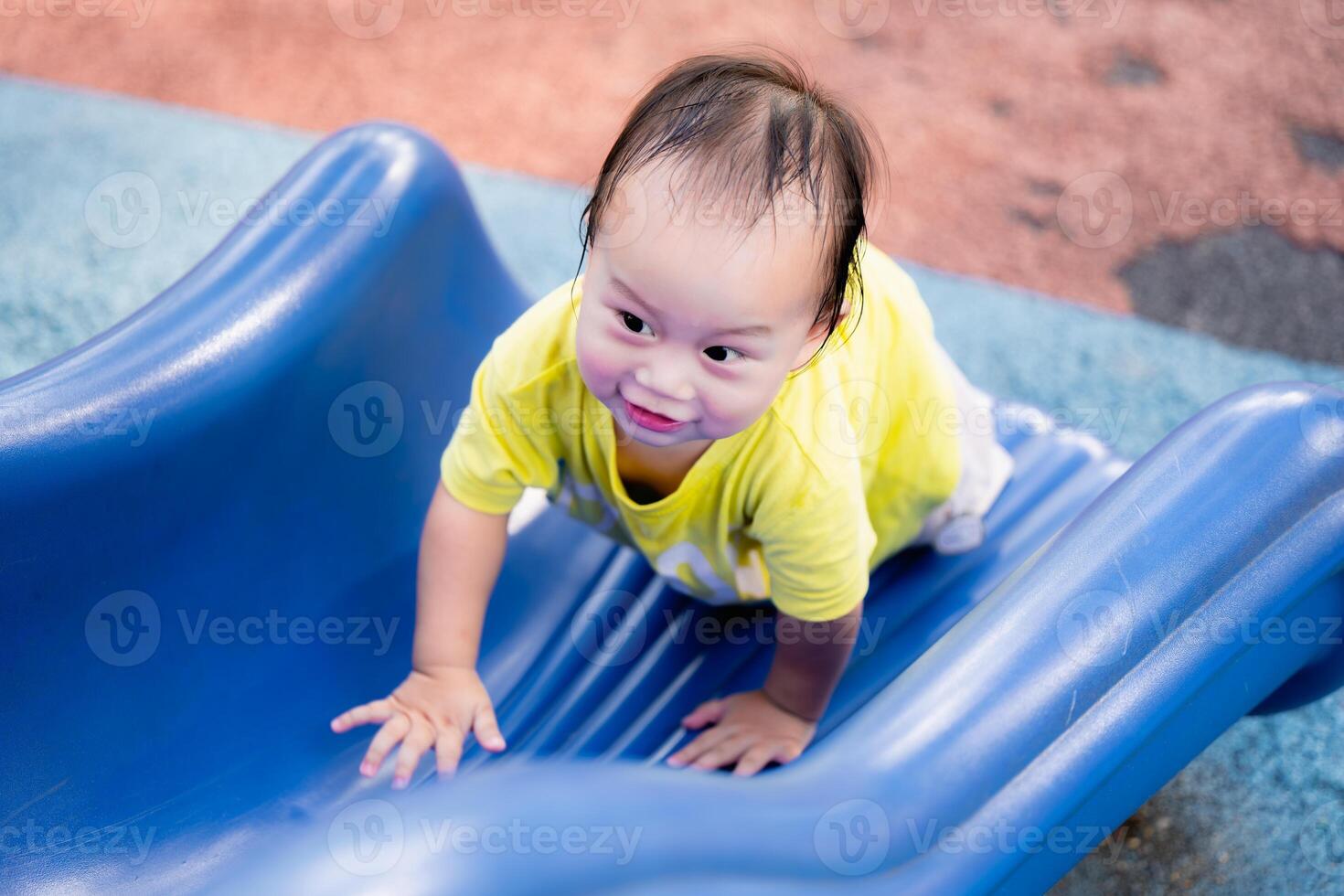 Portrait of Asian Baby Boy happily playing on the playground, and sliding down a blue slide. Cute kid climbs the slide on the playground. Toddler child aged one-year-old. Real People. photo