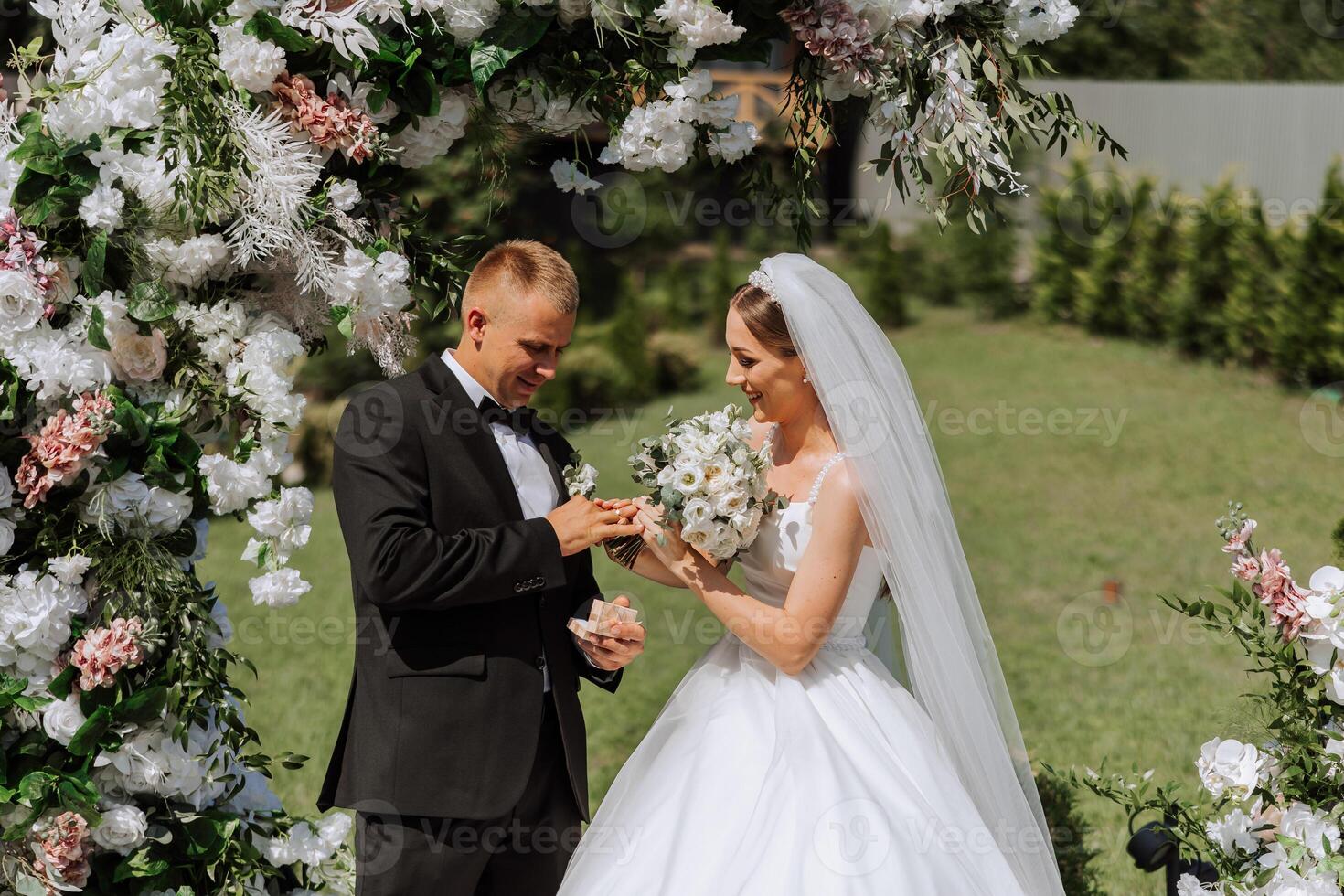 el novio pone en el de la novia Boda anillo durante el Boda ceremonia cerca el flor arco. verano Boda en naturaleza. ella dijo Sí. un conmovedor momento a el Boda ceremonia foto