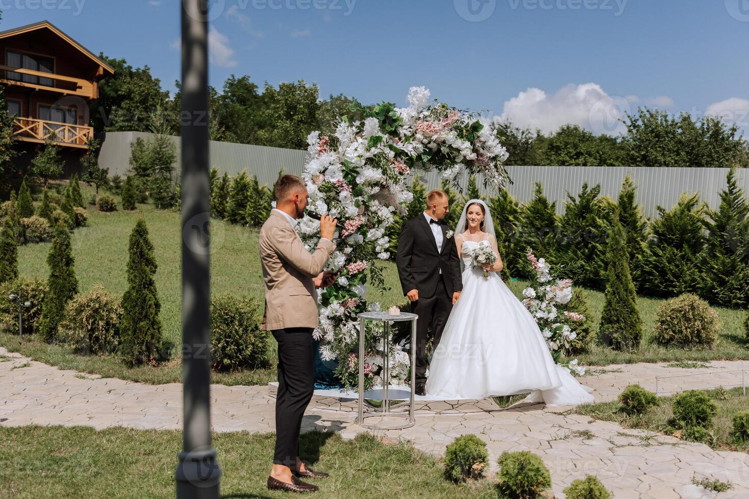 Wedding ceremony in nature. The bride and groom near the flower arch. The master of ceremonies at the wedding during the performance in the background of the bride and groom. photo