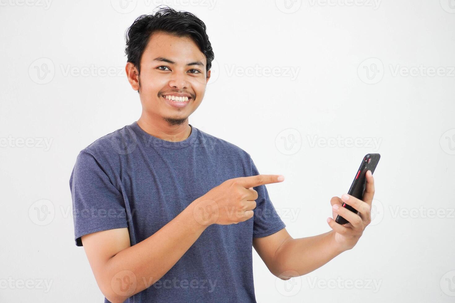 Excited smiling or happy young asian man in navy t-shirt holding phone and pointing at smartphone with finger, isolated on white background photo