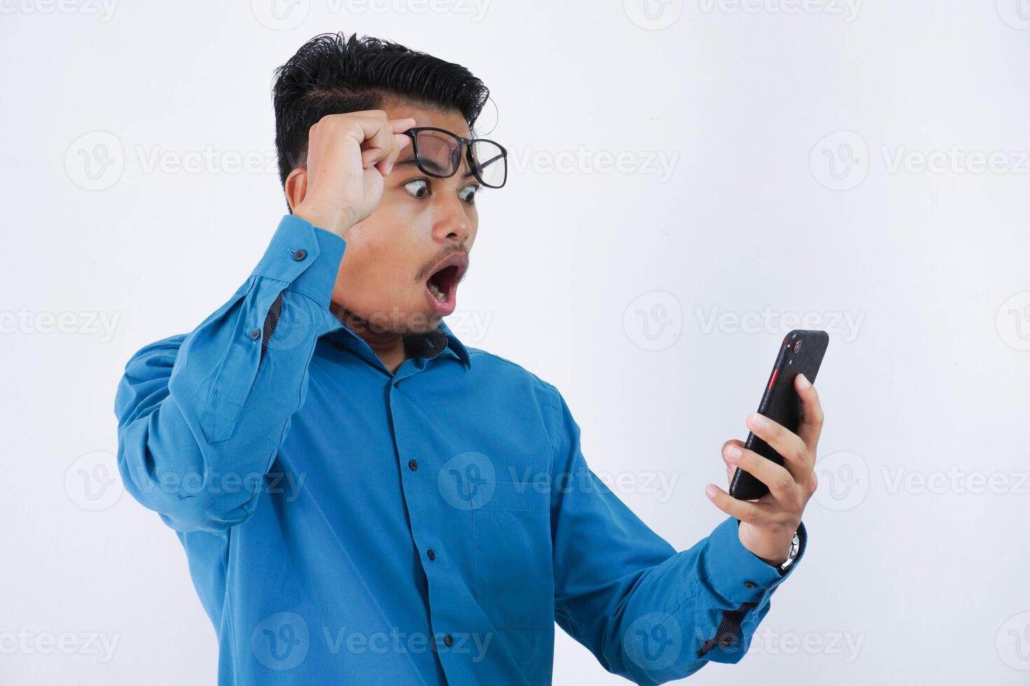 Portrait of surprised and shocked handsome young asian businessman with glasses in wearing blue shirt using smartphone and taking off glasses isolated on white background photo